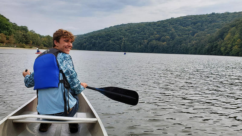 A smiling young man paddles a canoe on a lake.