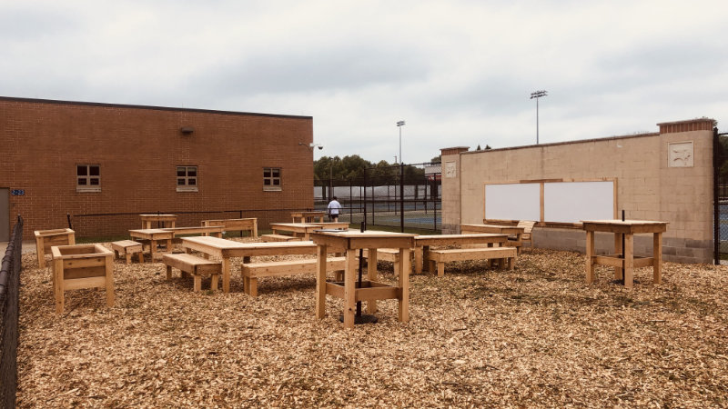 Simple wooden tables and benches sitting on wood chips next to a school.